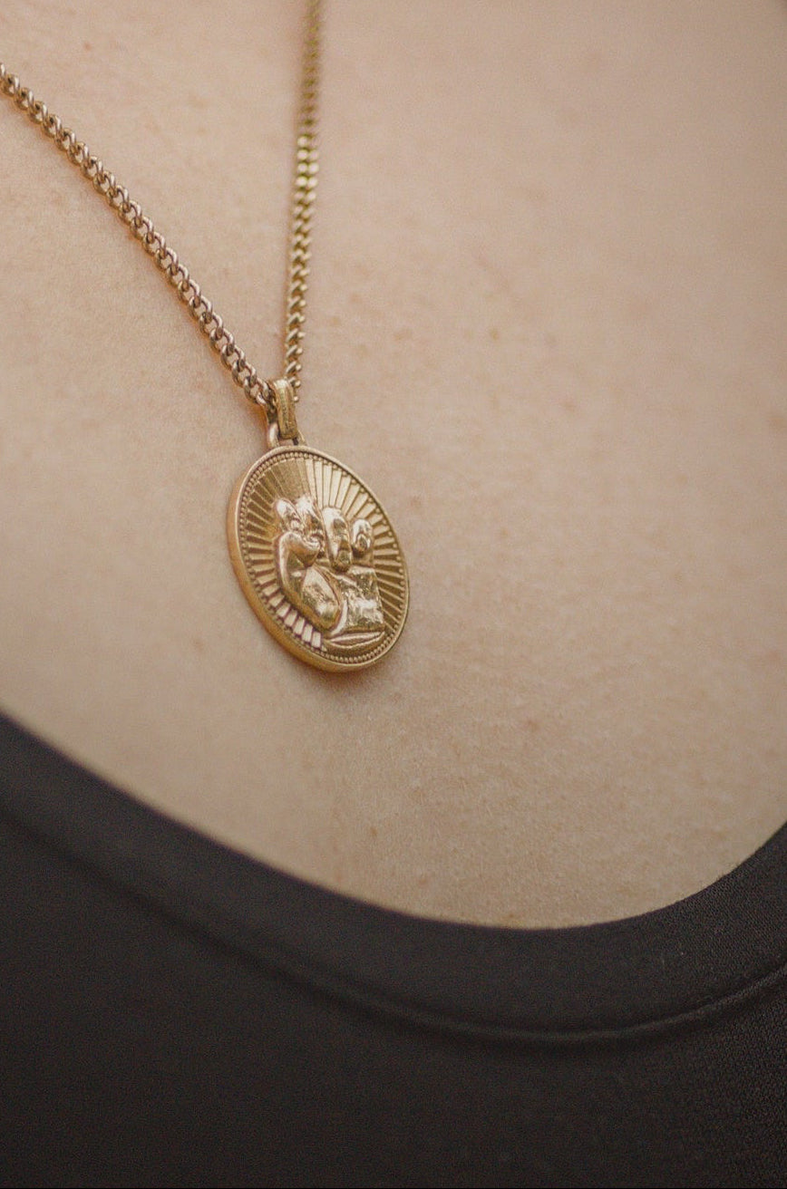 A gold pendant necklace with an oval sunburst design worn on a model's neck over a black top.